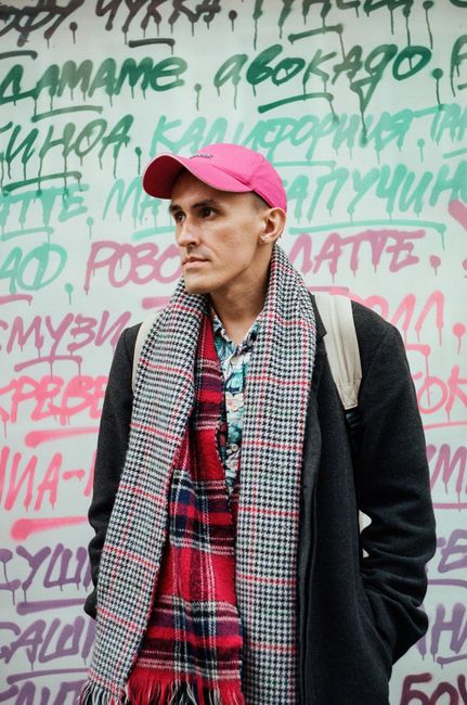 Trendy man in a pink cap and plaid scarf stands before a graffiti wall.