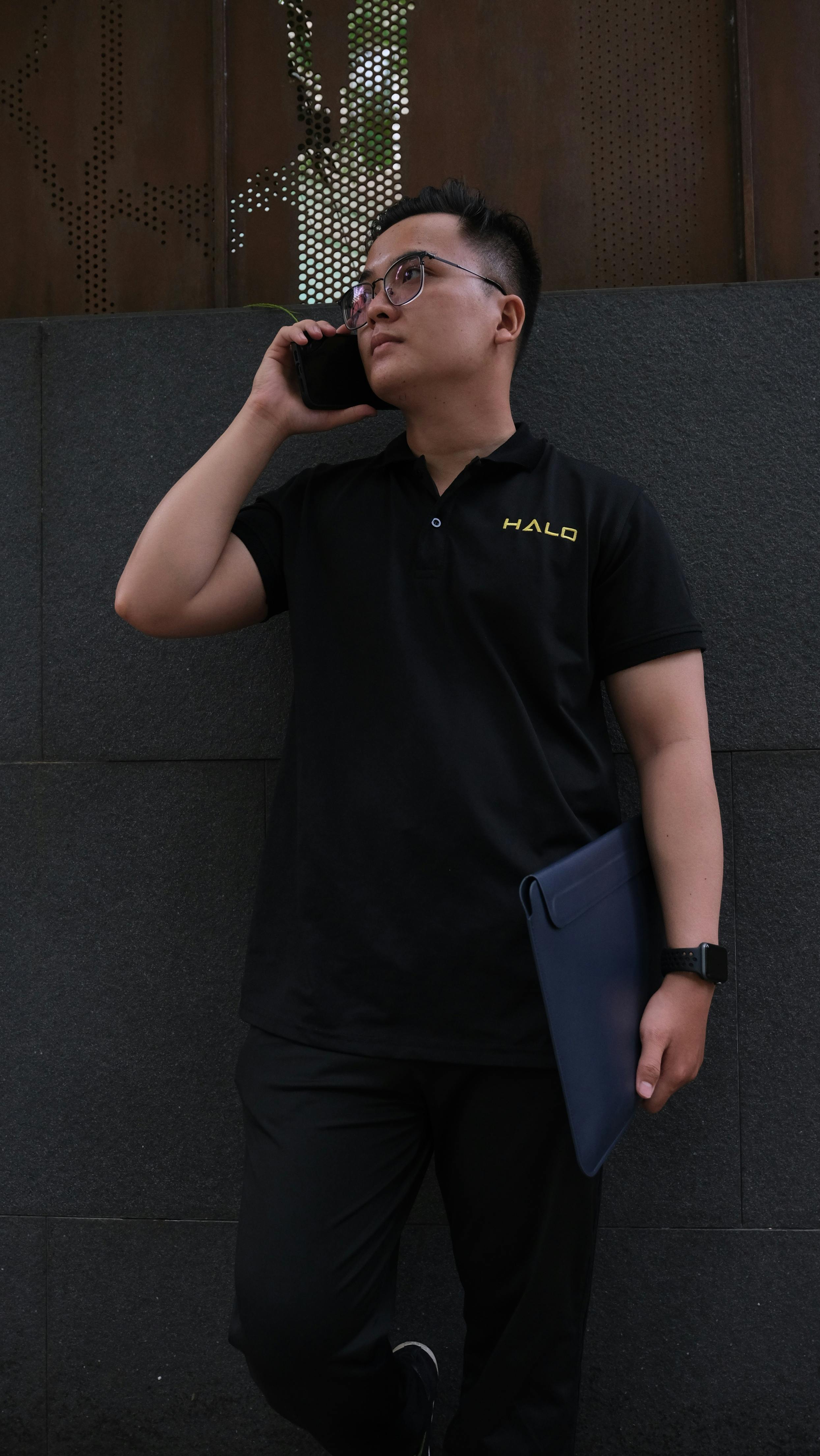 Young Asian man in polo shirt using smartphone and holding a folder against a city wall.