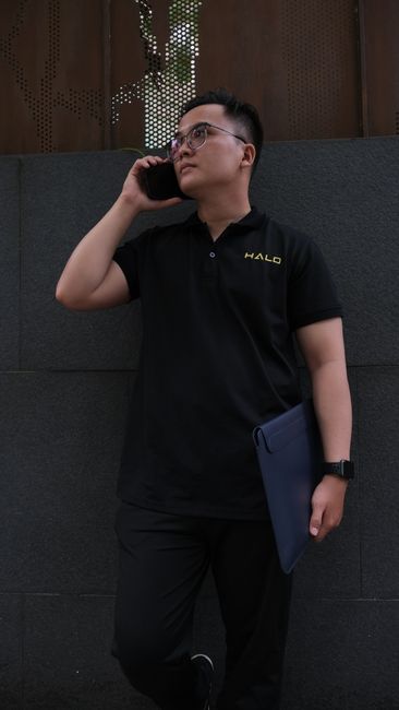 Young Asian man in polo shirt using smartphone and holding a folder against a city wall.