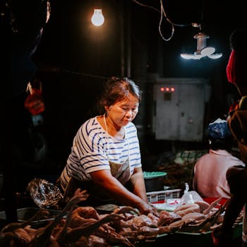 Asian woman vendor selling poultry under warm light at a night market. Captures the lively market atmosphere.