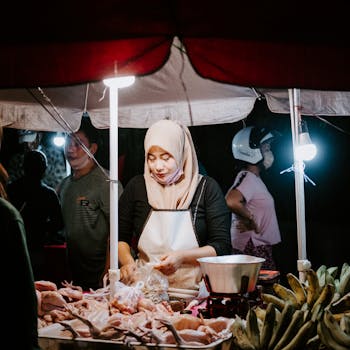 A Muslim woman in hijab works at a vibrant night market vendor stall, selling fresh produce.