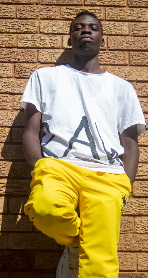 Free Young man posing in vibrant yellow pants against brick wall in sunny urban setting. Stock Photo