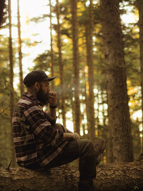 Free Contemplative man with beard smoking on a tree log in a serene forest setting. Stock Photo