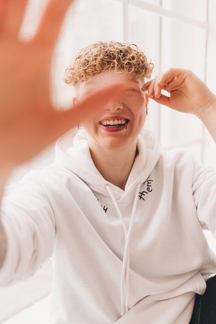 A cheerful person with curly hair laughs joyfully wearing a white hoodie indoors.