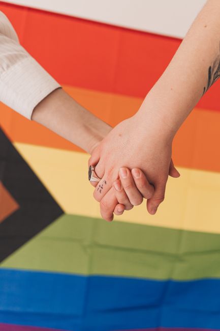 Two people holding hands in front of a vibrant progress pride flag, symbolizing unity and equality.