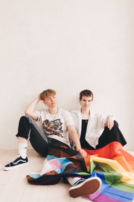 Two young adults sitting with a Progress Pride Flag in a studio setting.