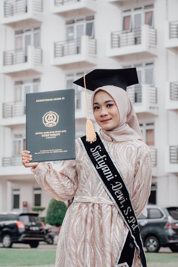 Young College Graduate Woman in Beige Hijab Holding Her Diploma and Standing with Graduation Cap