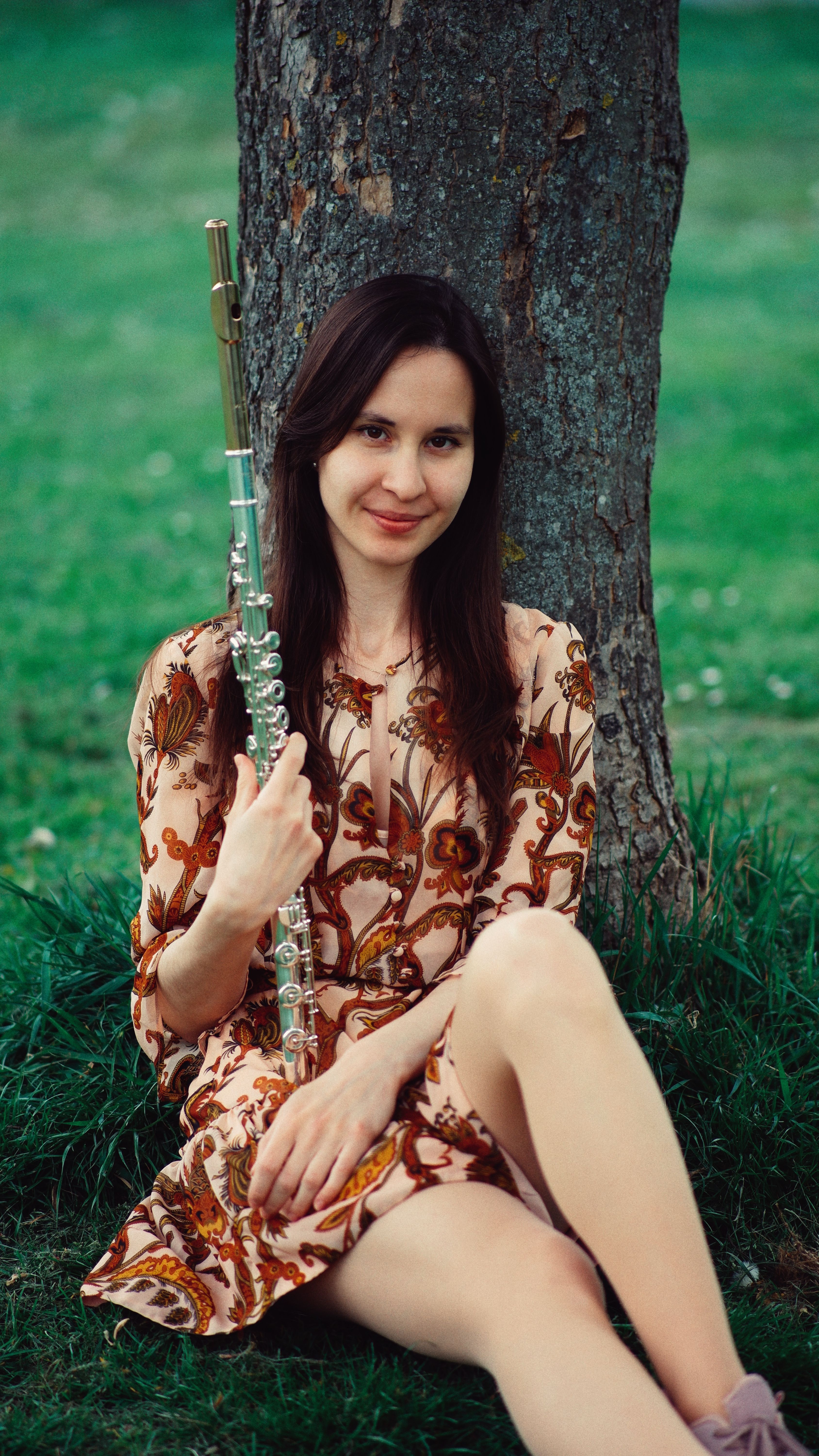 A woman holding a flute, sitting by a tree in a grassy field, smiling at the camera.