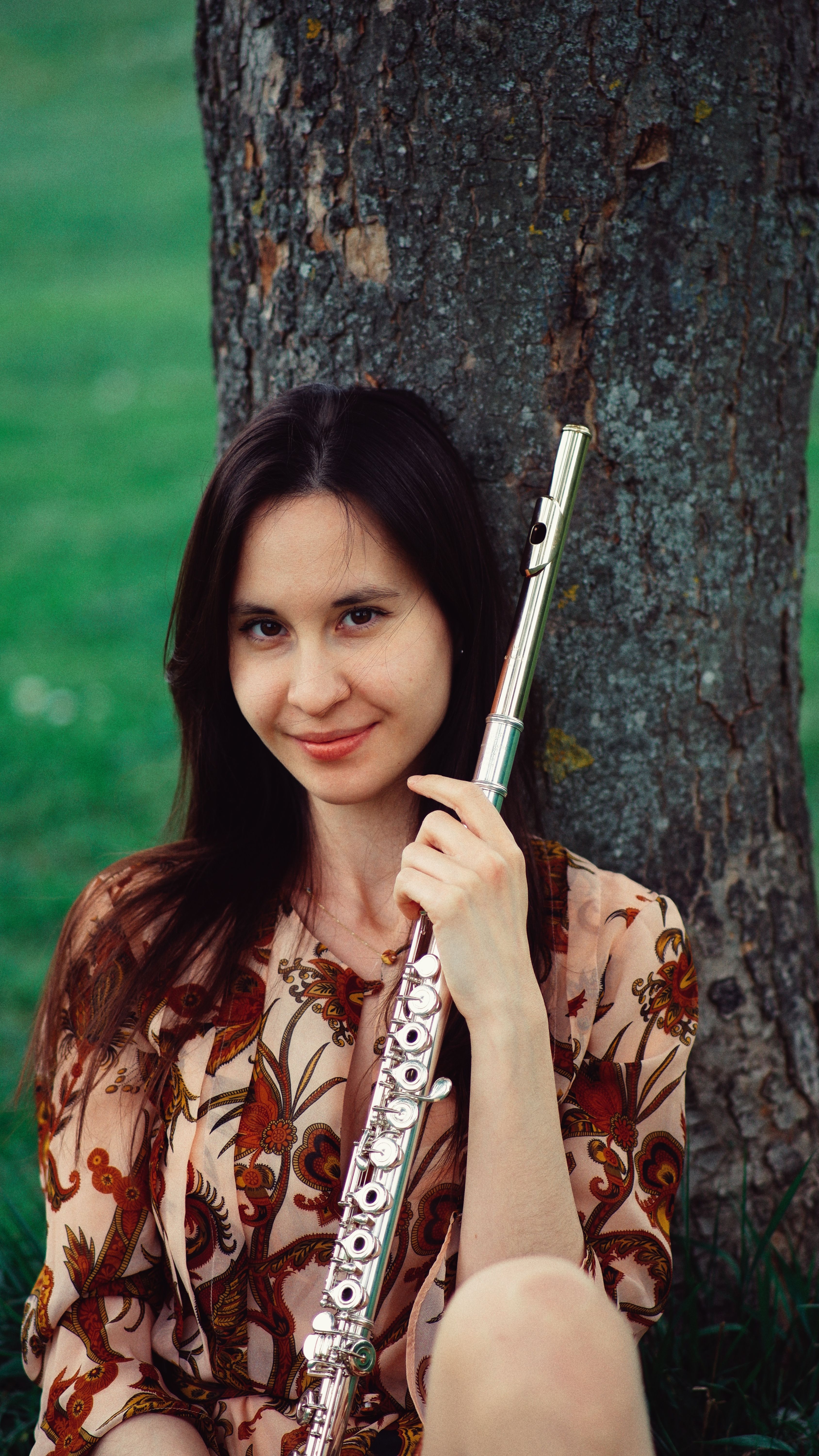 A woman smiles while holding a flute, seated against a tree in a park setting.