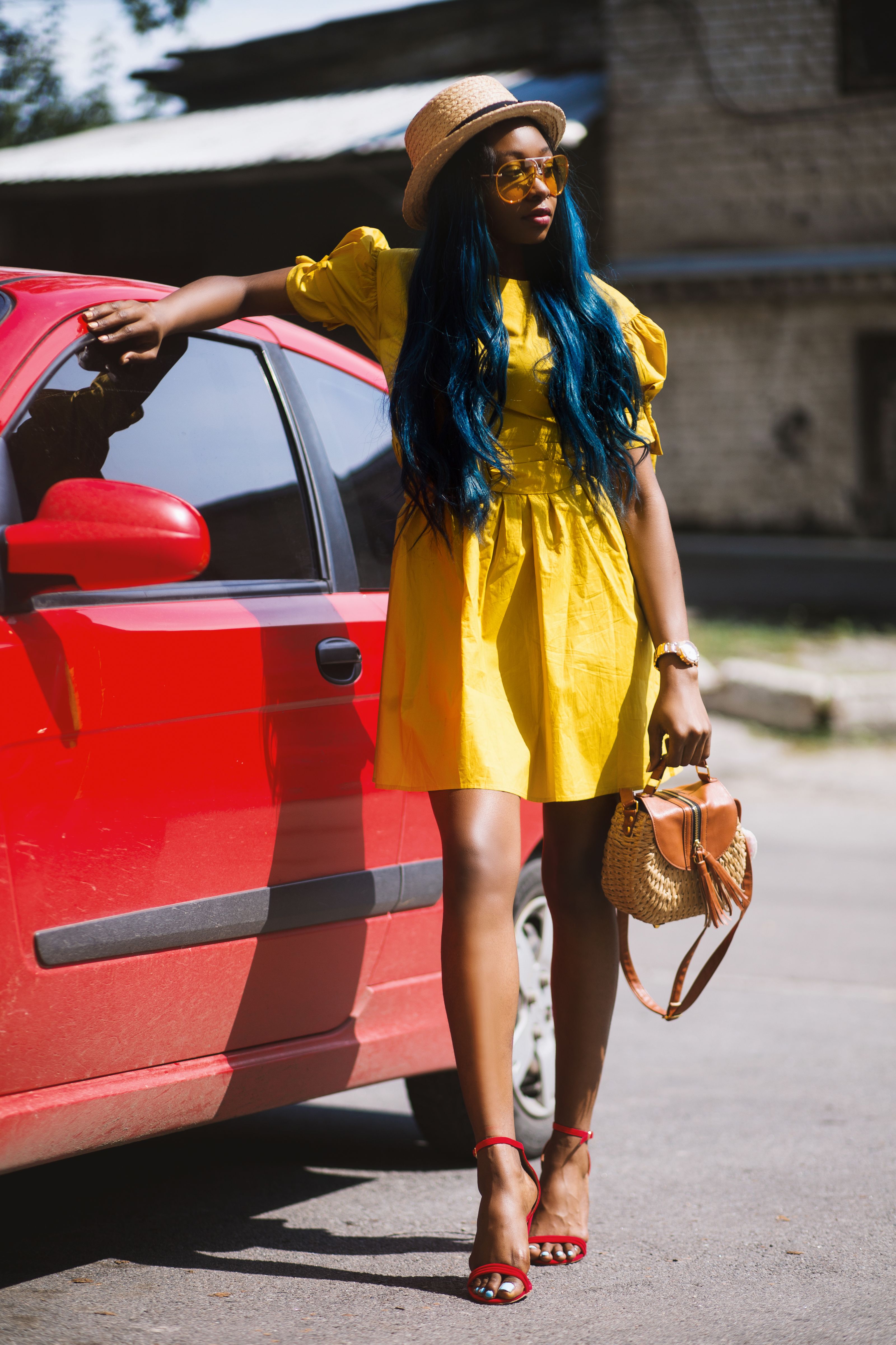 Mulher estilosa em vestido amarelo e saltos vermelhos posa ao lado de um carro em um dia ensolarado.
