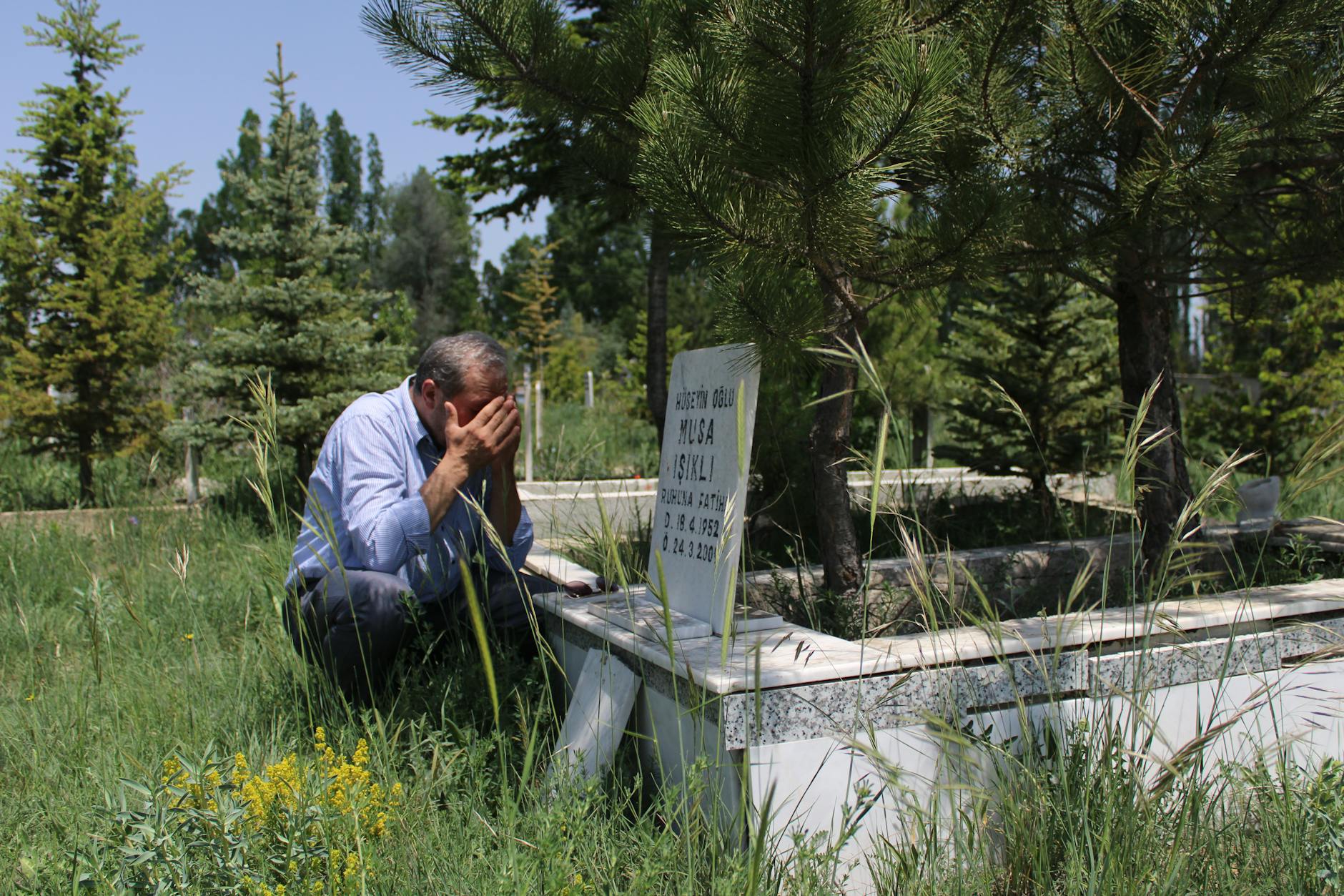 A man mourns at a grave, surrounded by lush greenery and serene cemetery.