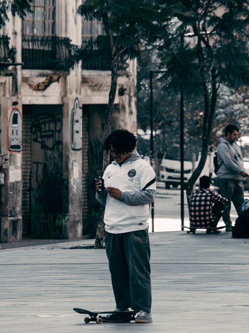 Free A young man with afro hair stands on a skateboard using his smartphone on an urban street. Stock Photo