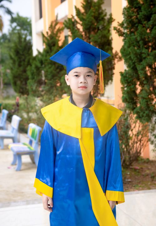 Free Young boy wearing a blue and yellow graduation gown smiling outdoors, symbolizing achievement. Stock Photo