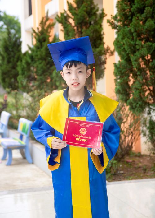 Free A young boy in a colorful graduation gown and cap proudly holding his diploma outdoors. Stock Photo