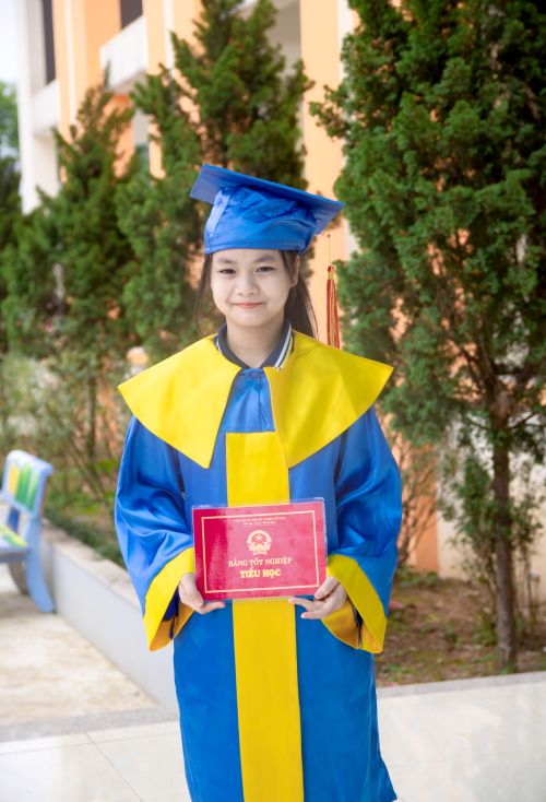 Free Cheerful schoolgirl in a colorful graduation gown holding diploma outdoors. Stock Photo