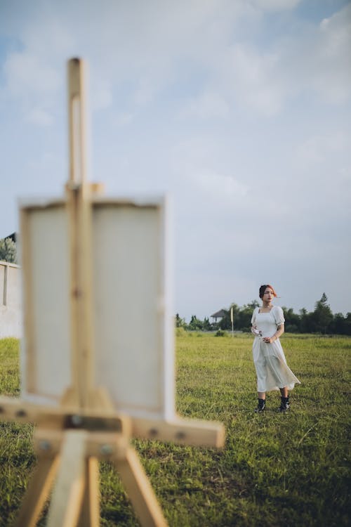 Woman in a White Dress Standing on a Grass Field with Canvas on an Easel 