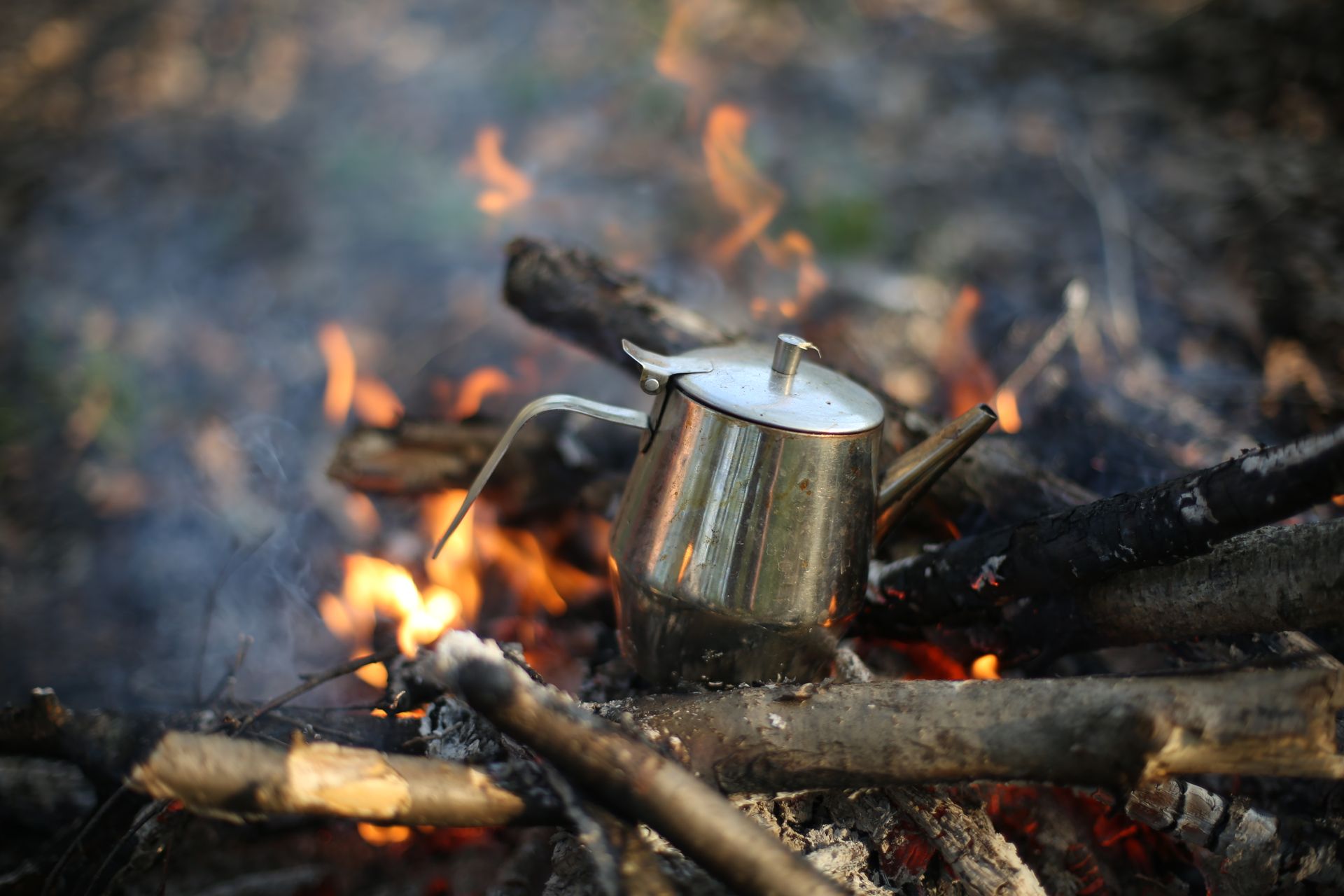 A stainless steel teapot heating over a campfire with burning wood outdoors. Perfect for camping themes.