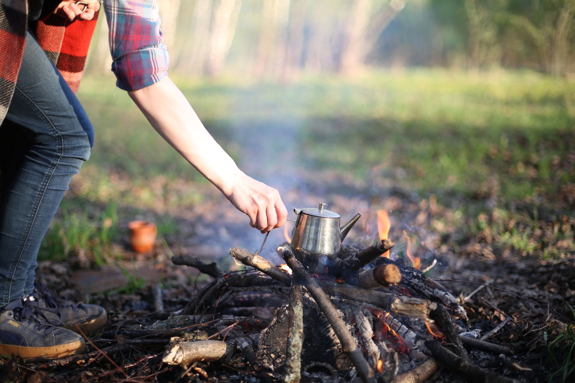 A person tends to a teapot heating over a campfire in a wooded area.