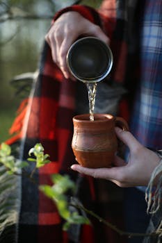 Close-up of water being poured into a clay mug outdoors surrounded by greenery.