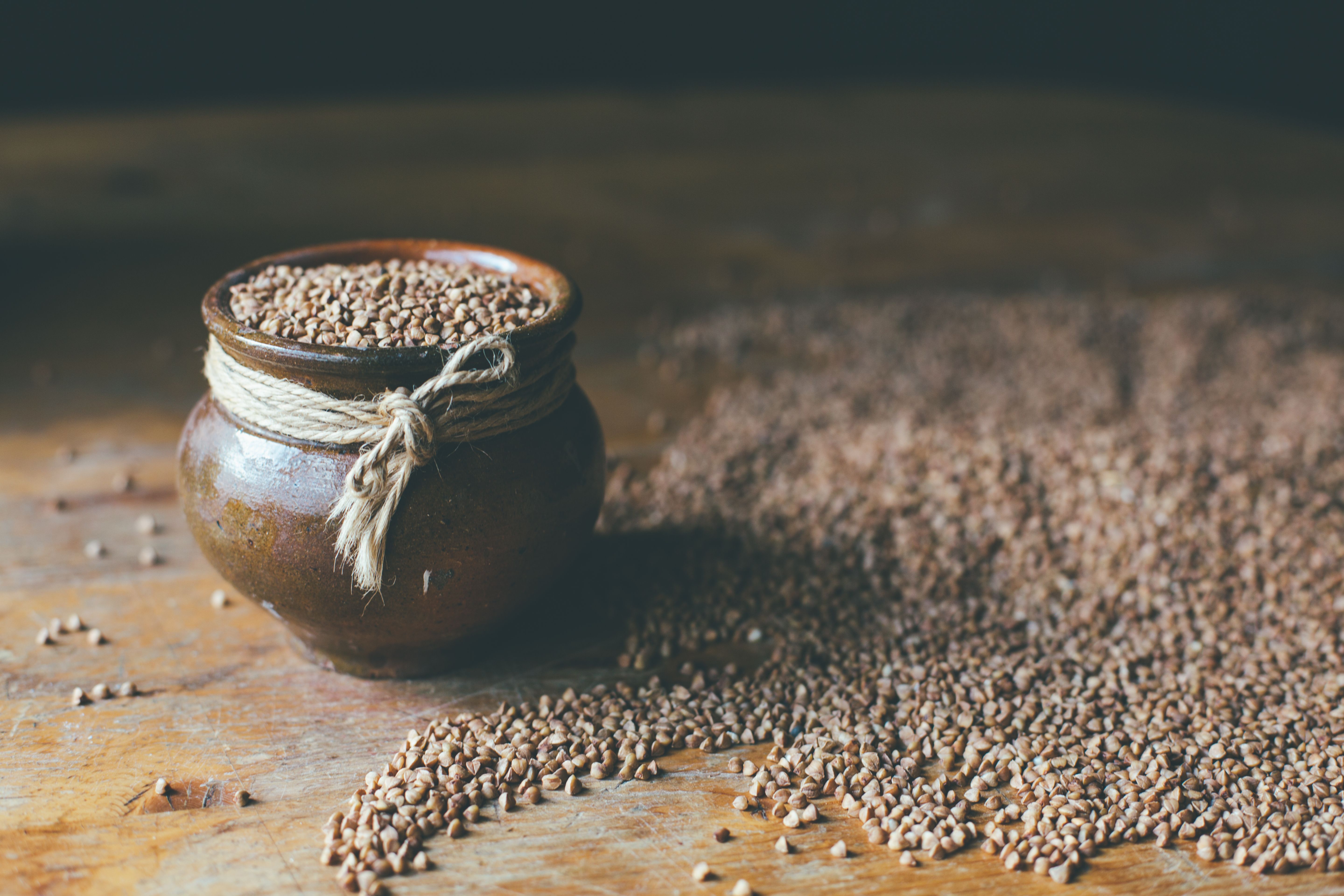 A rustic ceramic pot filled and surrounded by buckwheat grains on a wooden surface.