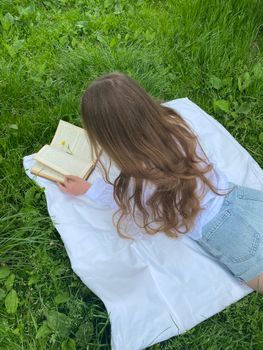 A woman in denim shorts and white top reading on a blanket in a grassy field.