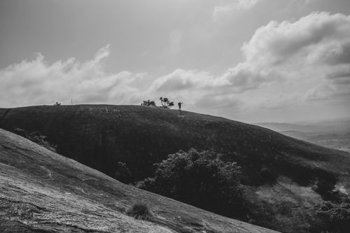 Free Black and white photo of a lone figure on Savandurga Hill with dramatic clouds and rugged terrain. Stock Photo
