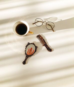 Flat lay of a vintage mirror, comb, cup of coffee, and eyeglasses on a sunlit surface.