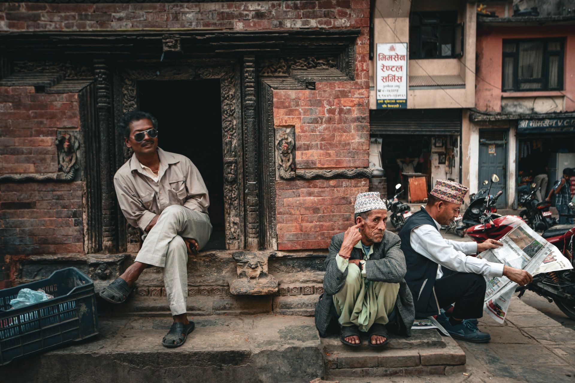 Three men sitting and reading newspapers on a historic street in Nepal, capturing a candid moment.
