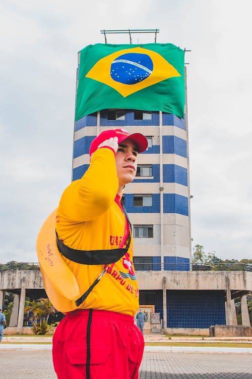 Man in Yellow Hoodie Standing in Front if Building with Brazilian Flag Displayed