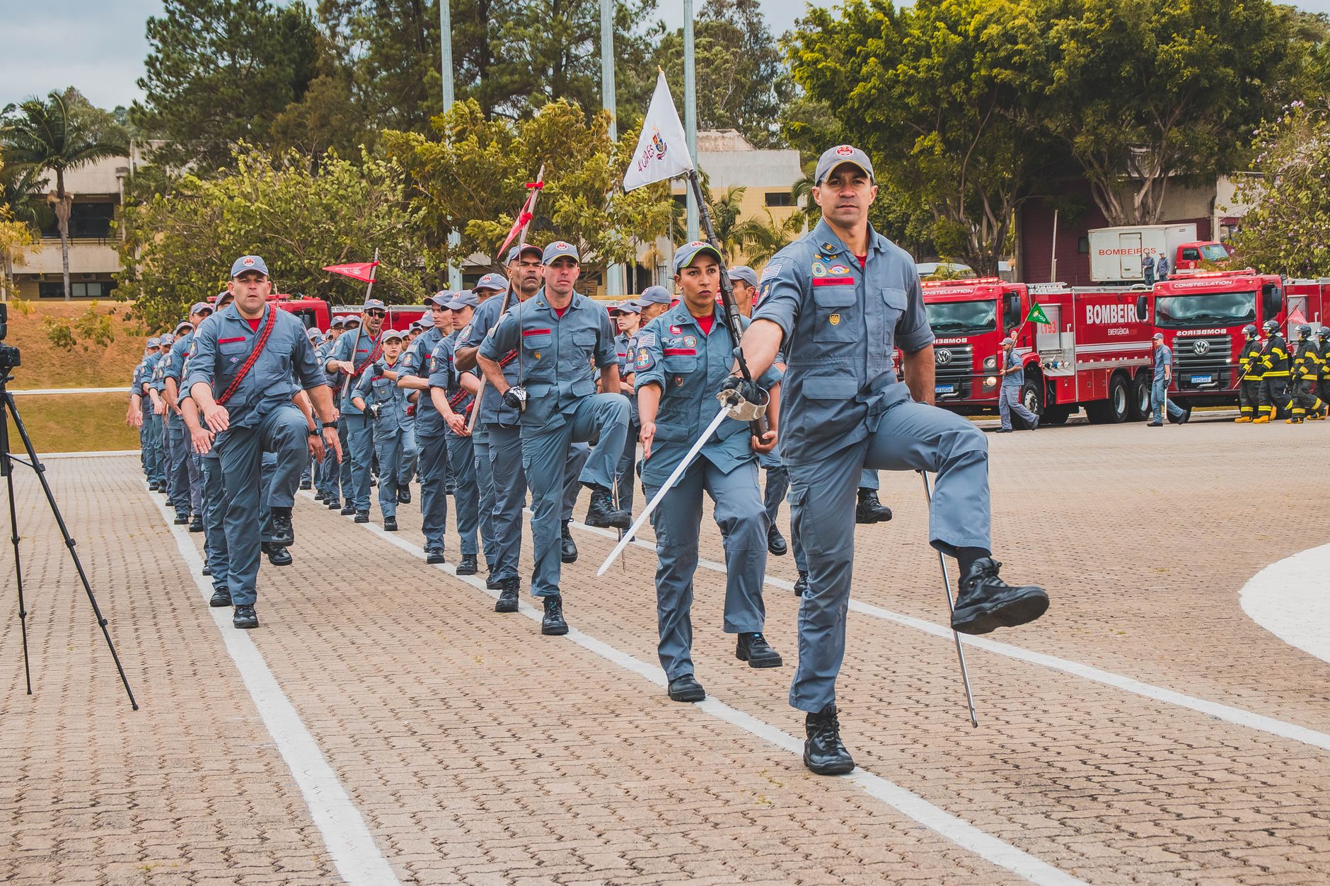 Soldiers in uniform march in a ceremonial parade with flags and fire trucks in the background.