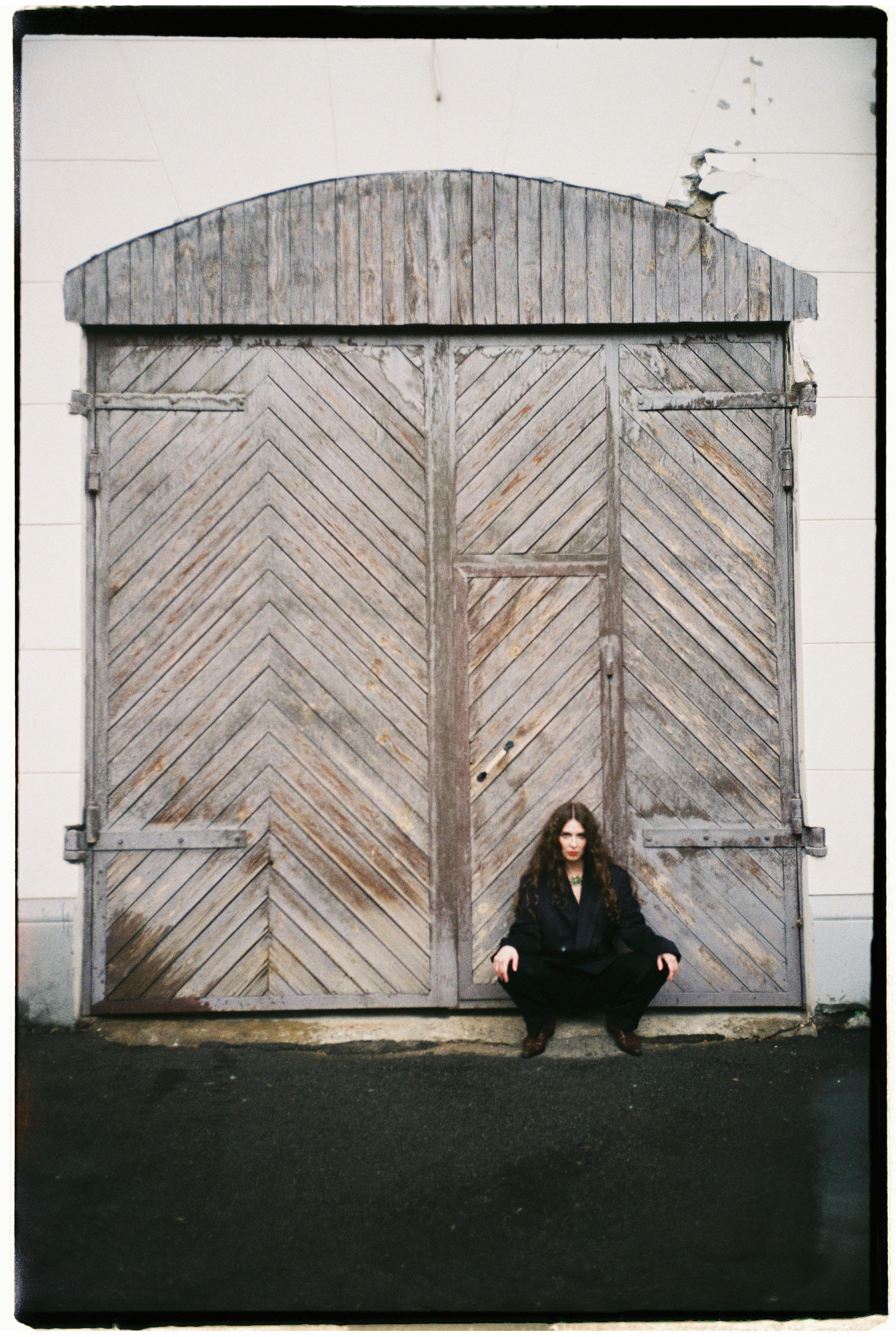 A woman in black clothing sits in front of a large rustic wooden door, creating a moody atmosphere.