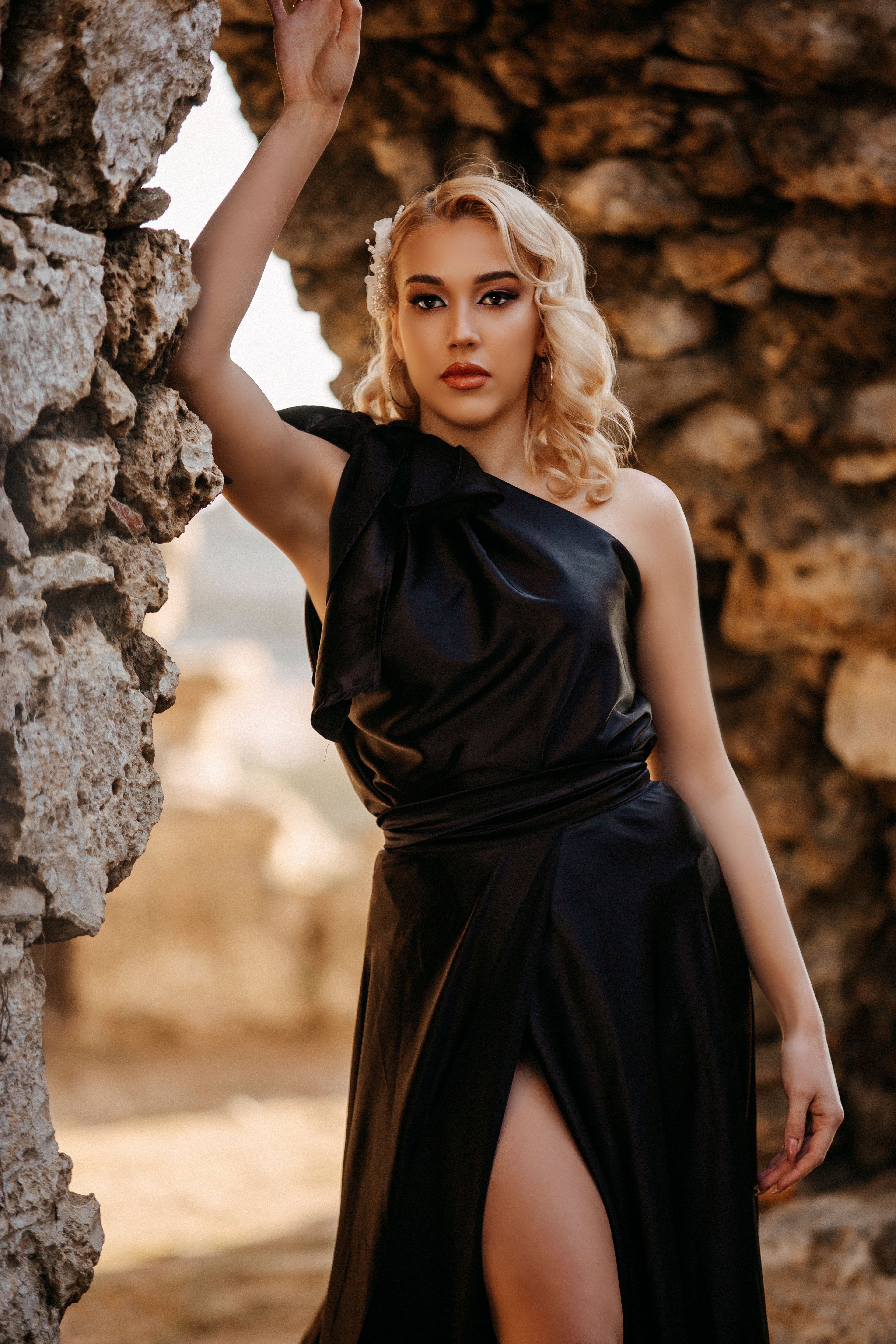 A stylish woman poses elegantly in a black dress against a stone archway, exuding confidence and grace.