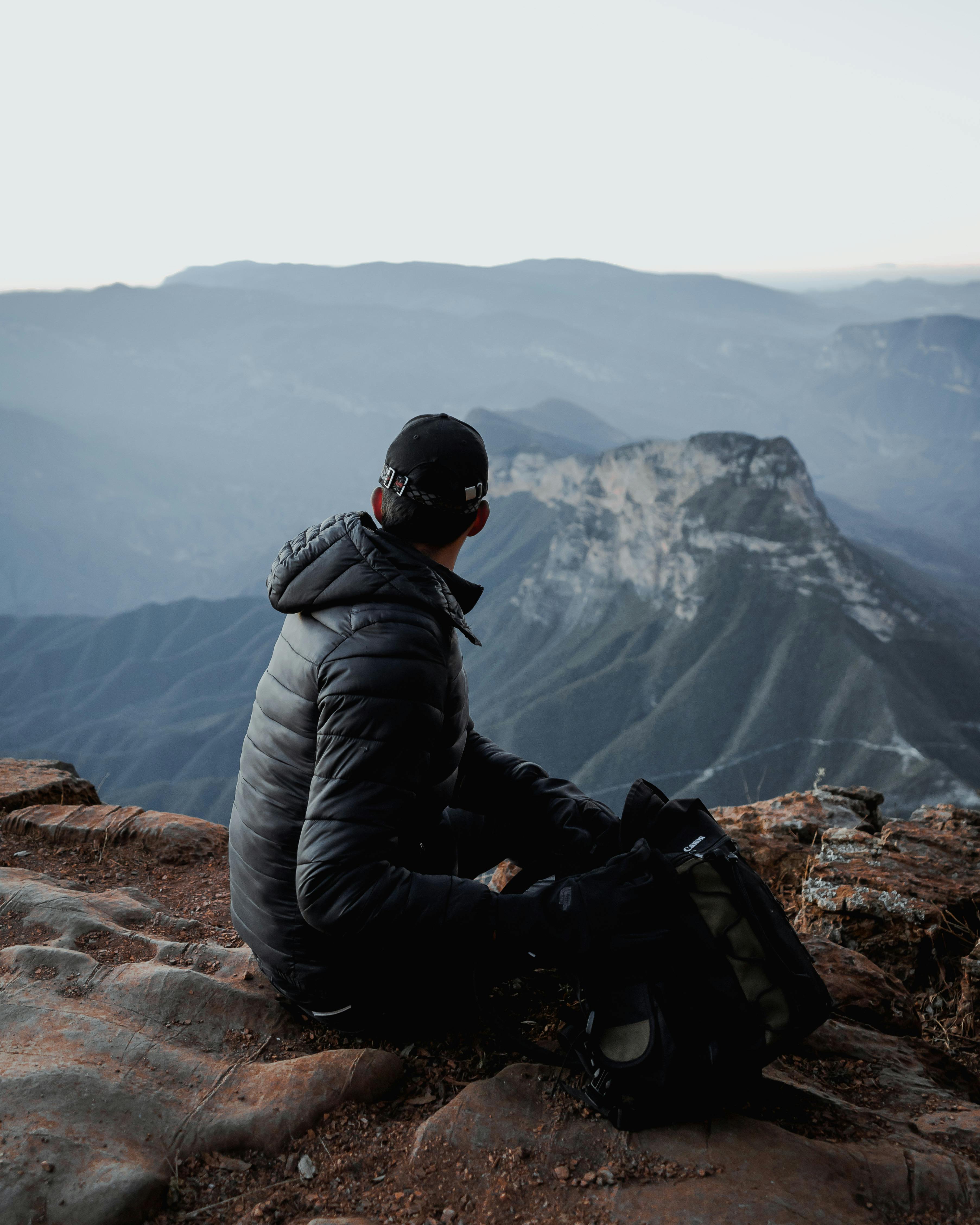 man in a jacket sitting at the top of a mountain and looking at view