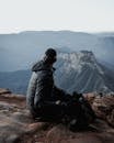 Man in Black Jacket and Black Helmet Sitting on Rock Looking at Mountains