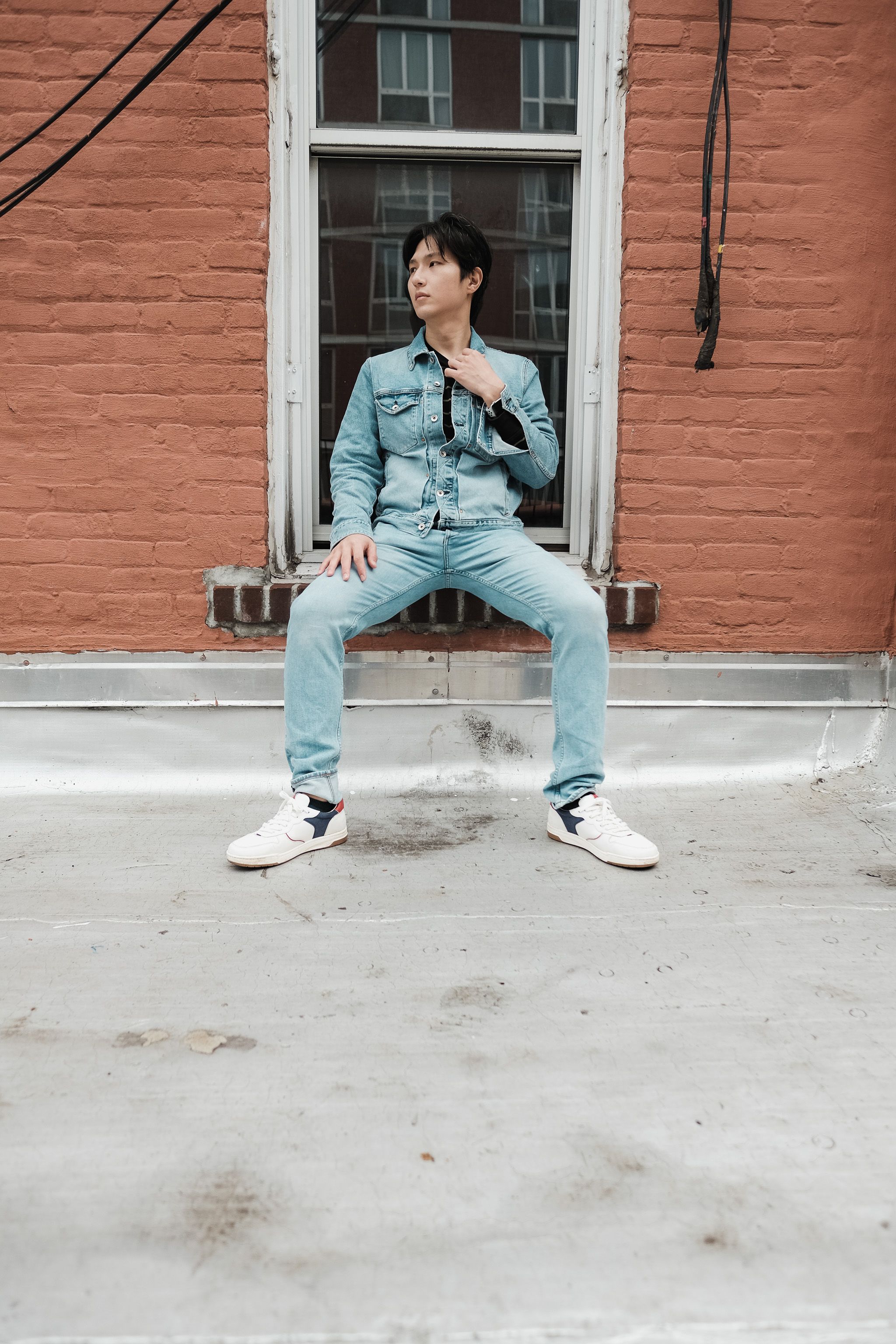 Stylish young man in denim outfit sitting on a window sill, showcasing urban fashion.