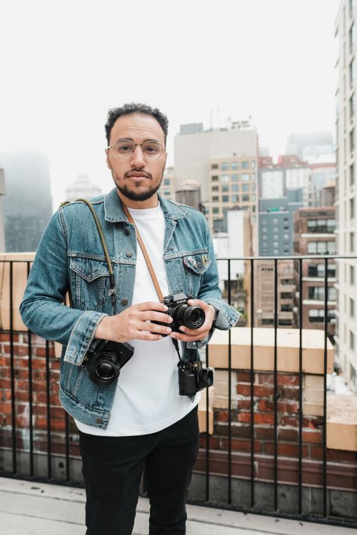 Free Man in denim jacket holding cameras on a city rooftop with tall buildings in the background. Stock Photo