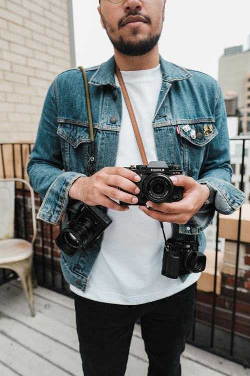 Free Man holding multiple cameras on a rooftop, wearing a denim jacket and white shirt. Stock Photo