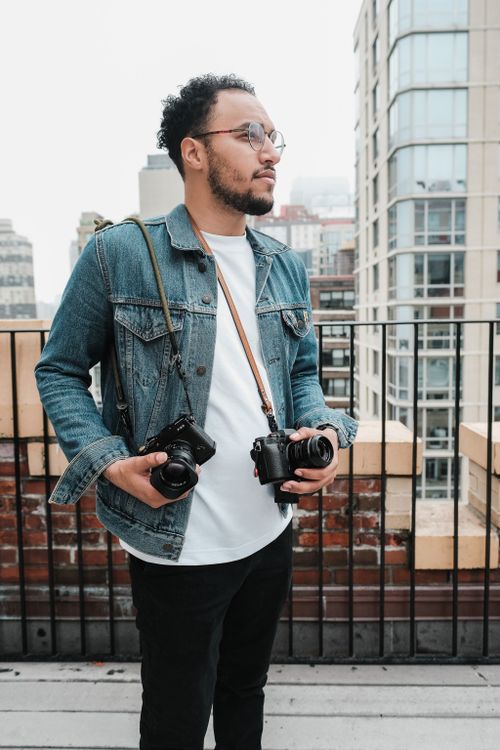 Free Man in denim jacket stands on rooftop with cameras, amidst a cityscape background. Stock Photo