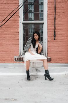 Stylish woman in a dress and boots posing on a rooftop windowsill for a modern fashion shoot.