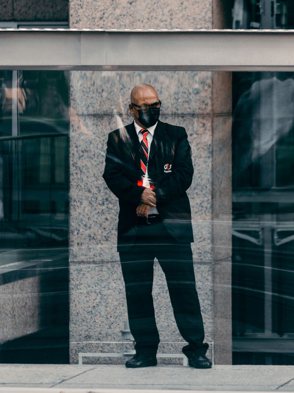 Photo by Anderson Santos Bald security guard in black suit and tie stands in an urban environment, providing security.