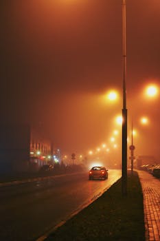 A foggy urban street illuminated by streetlights and a lone car in the night.