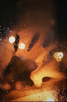 A mysterious handprint on a rain-covered glass window illuminated by warm bokeh lights at night.
