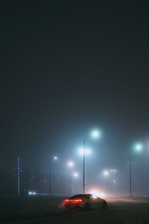 Free A car navigates through a foggy street illuminated by street lights, creating a mysterious night scene. Stock Photo