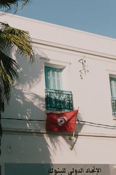 A Tunisian flag displayed on a white facade with turquoise windows and ornate iron balconies.