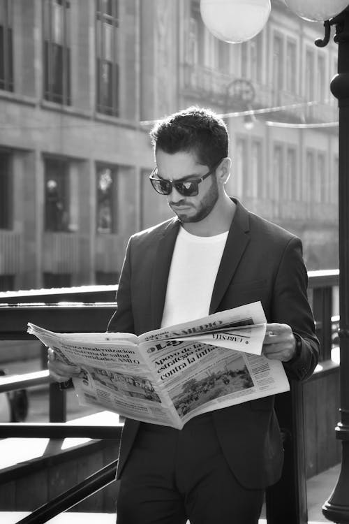 Grayscale Photo of a Man in Business Suit Wearing Sunglasses while Holding a News Paper