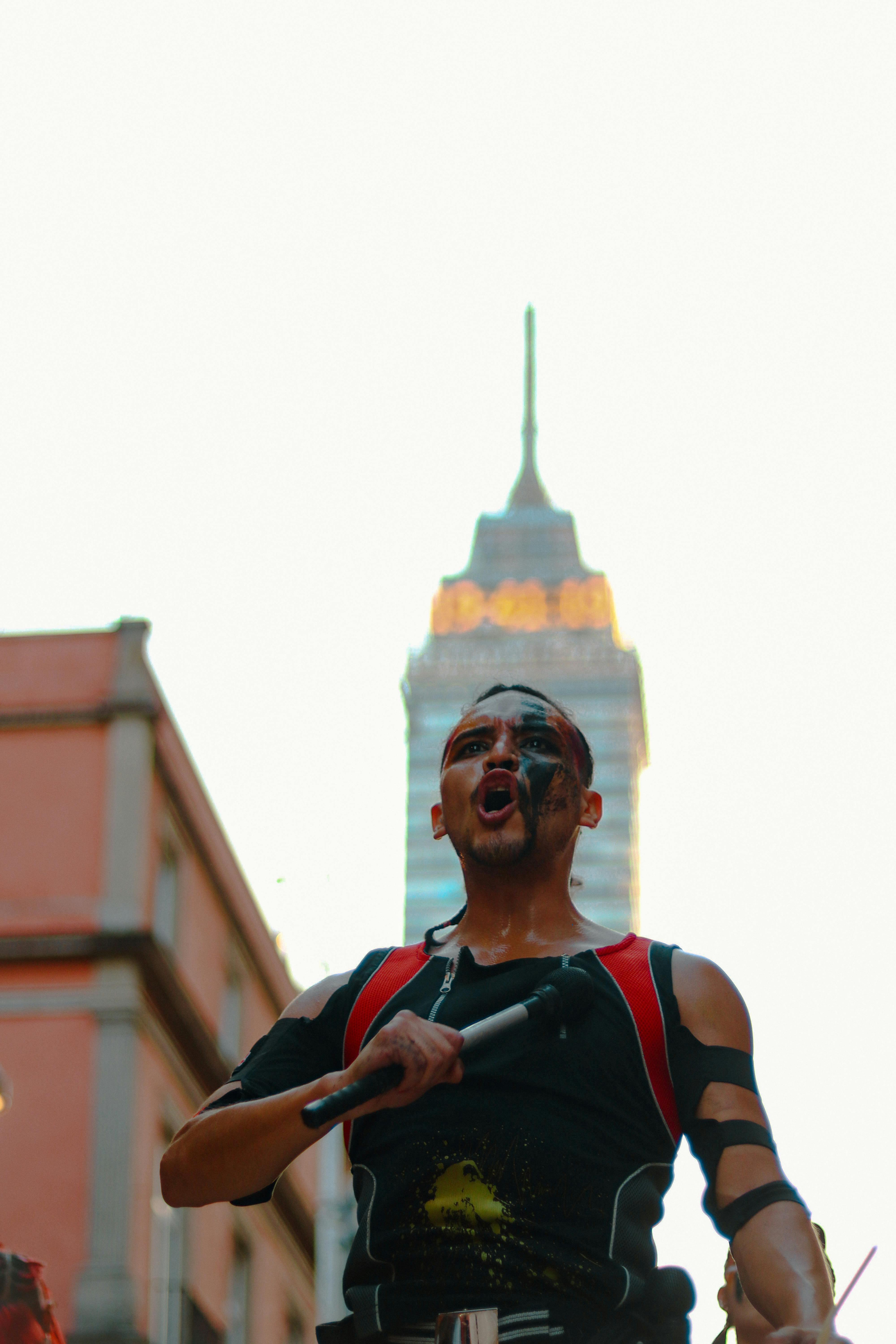 Energetic performer with face paint at a vibrant parade in Mexico City. Iconic tower in background.