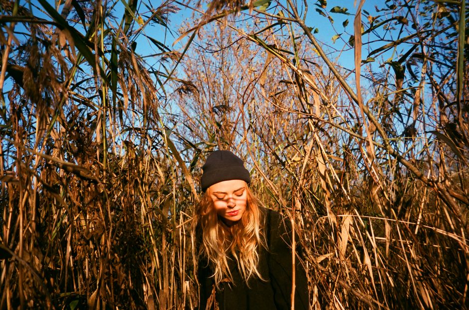 A young woman wearing a beanie stands amidst tall autumn reeds in a rural field