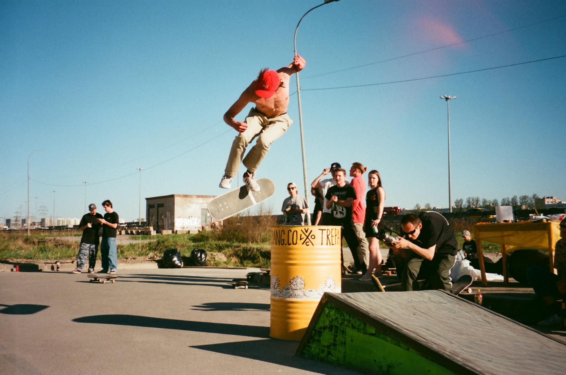 Skateboarder performs an aerial trick over a barrel in an urban park setting, surrounded by an audience.
