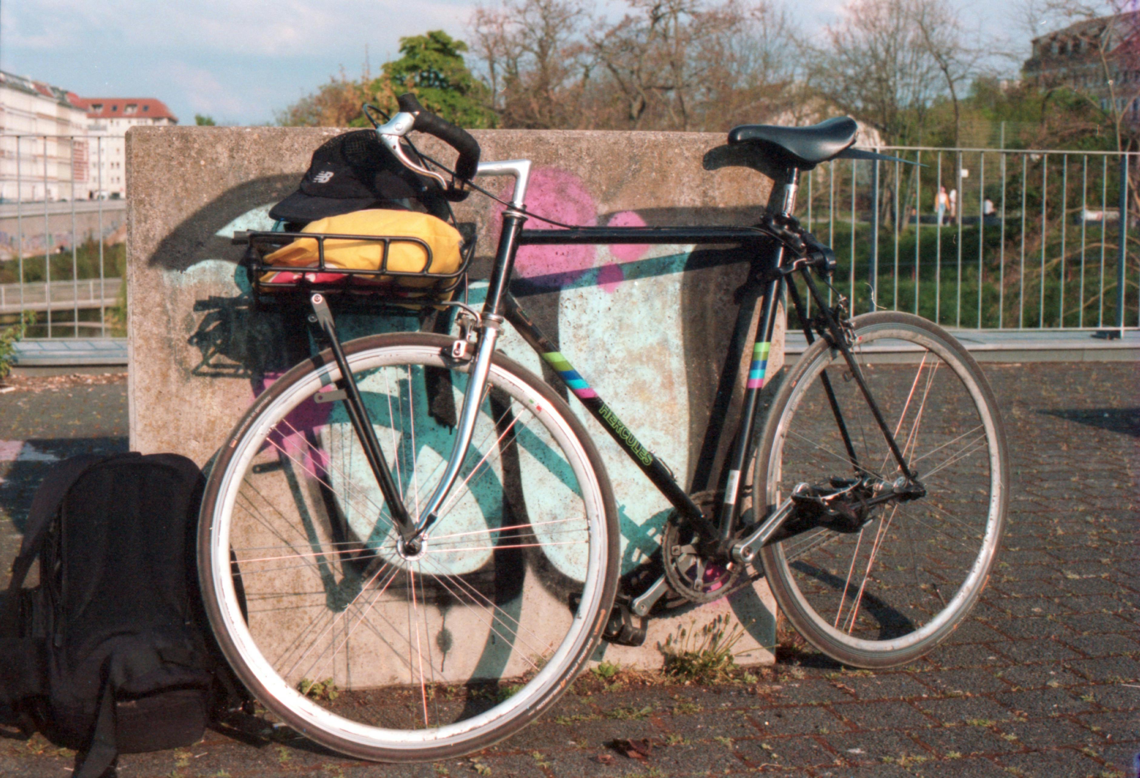 A vintage bicycle parked outdoors against a graffiti-covered wall, with a backpack nearby.