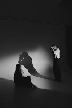 Artistic black and white photo capturing a female photographer shooting a seated subject in a studio.